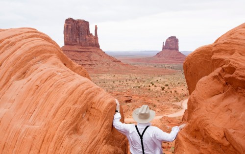 A person in a white shirt and hat stands between red rock formations, overlooking a desert landscape with distant mesas.