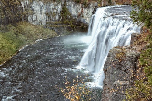 A cascading waterfall flows into a river, surrounded by rocky cliffs and lush greenery.