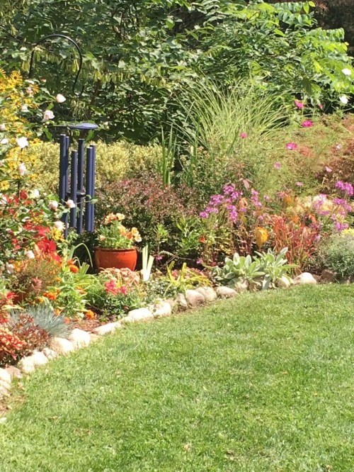 A vibrant garden with colorful flowers, greenery, and a wind chime, bordered by a stone path and lush grass.