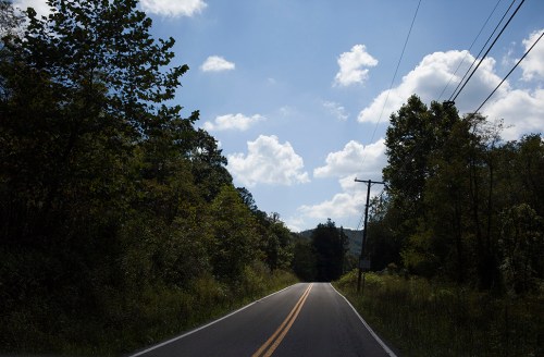 A winding road stretches into the distance, flanked by trees under a blue sky with fluffy clouds.