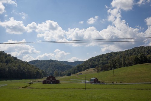 Two red barns sit in a green valley, surrounded by rolling hills and a blue sky with fluffy white clouds.