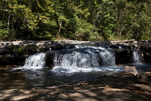 A serene waterfall cascades over rocks, surrounded by lush green trees in a peaceful natural setting.