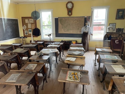 A vintage classroom with wooden desks, a map on the wall, and a mannequin dressed as a teacher.