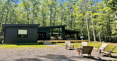 wooden modern cabin with large trees in the back and a small outdoor table and chair set in the front on a sunny day