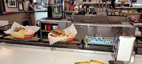 Two baskets of food with burgers and fries on a counter, with a toy train carrying a drink in the background.