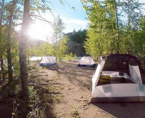 A serene campsite by a river, featuring several tents surrounded by trees and sunlight filtering through the leaves.