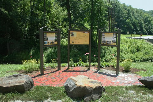 Three informational signs on a brick path surrounded by greenery and rocks, near a road in a natural setting.
