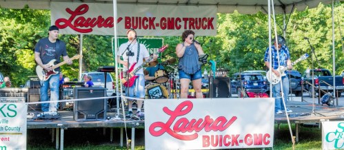 A band performs on stage under a tent, with a banner displaying "Laura BUICK-GMC" in the background.