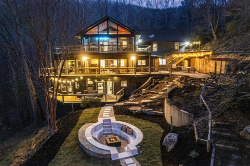 A modern mountain house at dusk, featuring multiple decks, warm lights, and a stone fire pit in the foreground.