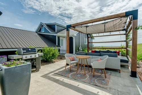 A cozy outdoor patio with a seating area, table, grill, and a pergola, surrounded by greenery and a scenic view.