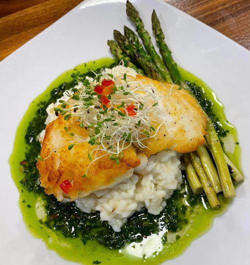 A plate of risotto topped with crispy fish, drizzled with green sauce, and served with asparagus and microgreens.