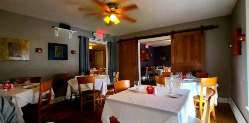 Cozy restaurant interior with white tablecloths, wooden accents, and warm lighting, featuring empty tables and a welcoming atmosphere.