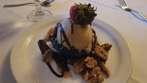 A dessert featuring a scoop of ice cream topped with a strawberry, drizzled with chocolate, and surrounded by nuts.
