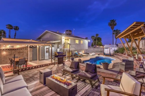 A backyard at dusk featuring a pool, lounge chairs, and a patio with string lights and palm trees in the background.