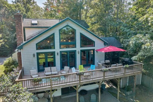 Aerial view of a spacious house with a large deck, outdoor seating, and a red umbrella, surrounded by trees.