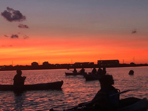 Silhouettes of kayakers on a calm waterway at sunset, with vibrant orange and purple hues in the sky.