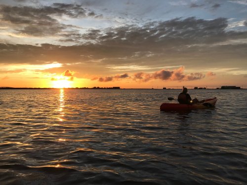 A person kayaking on calm water at sunset, with vibrant colors reflecting on the surface.