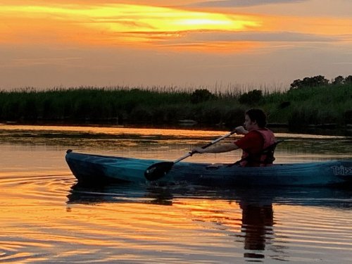 A person paddles a kayak on calm water during a vibrant sunset, with lush greenery in the background.