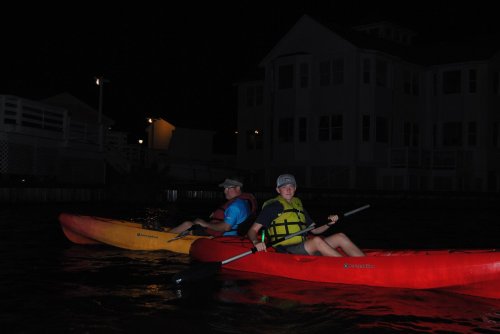 Two people kayaking at night, one in a yellow kayak and the other in a red kayak, with a house illuminated in the background.