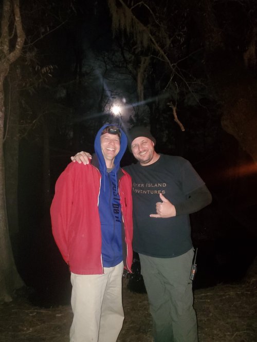 Two men stand together outdoors at night, smiling under a bright moon, surrounded by trees. One is wearing a red jacket.
