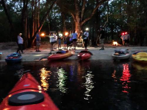 A group of people on a beach at dusk, with kayaks in the foreground and lights illuminating the gathering.