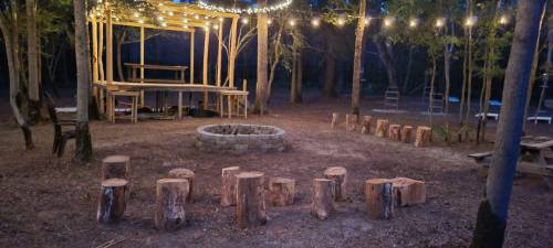 A cozy outdoor space with a wooden stage, string lights, and a circular arrangement of log seating around a fire pit.