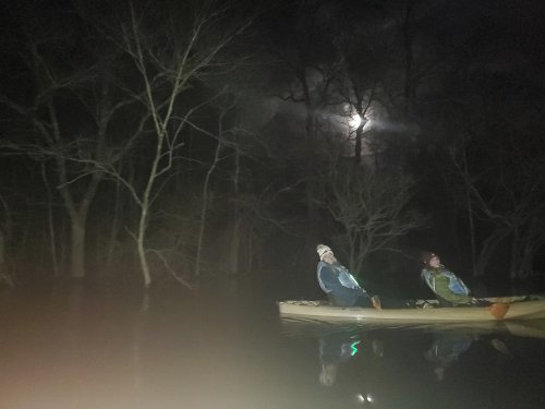 Two people in kayaks on a dark, flooded river, illuminated by moonlight and surrounded by bare trees.