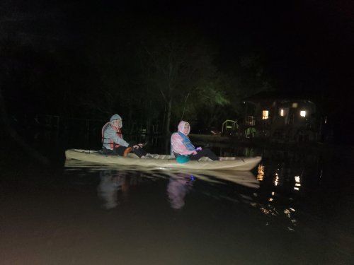 Two people in a kayak at night, paddling on calm water near a house with lights in the background.