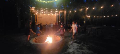A group of people gather around a fire pit at night, with string lights illuminating the outdoor space.