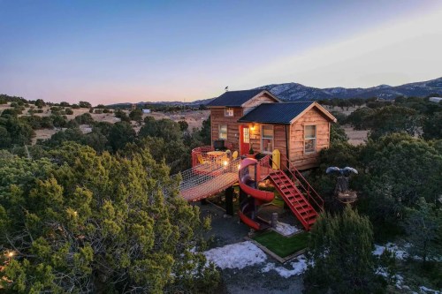 A wooden treehouse with a slide and stairs, surrounded by trees and mountains at sunset.