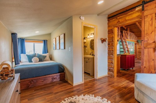 Cozy bedroom with a blue bedspread, wooden accents, and a bathroom, featuring natural light and warm decor.