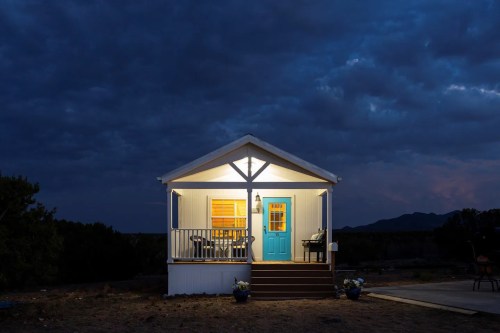 A cozy house illuminated at night, featuring a blue door and porch with chairs, set against a cloudy sky.