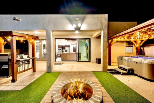 A modern outdoor space at night featuring a fire pit, patio, and hot tub, with cozy lighting and a view of the kitchen.
