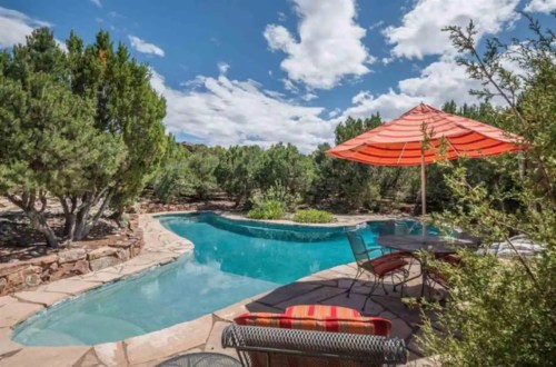 A serene pool area surrounded by greenery, featuring a shaded table under an orange umbrella and a clear blue sky.