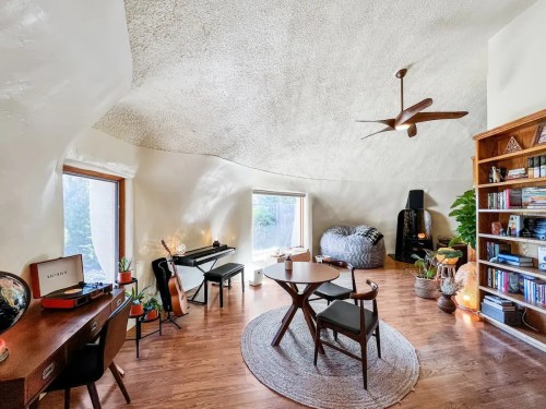Cozy room with a curved ceiling, wooden floor, plants, a bookshelf, and a table with chairs, featuring natural light.