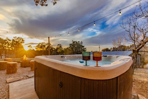 A hot tub with colorful drinks sits outdoors, surrounded by trees and a sunset, with string lights overhead.