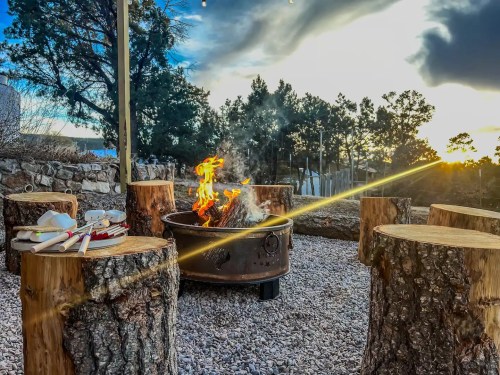 A cozy fire pit surrounded by log seating, with a sunset and trees in the background. Warm flames flicker.