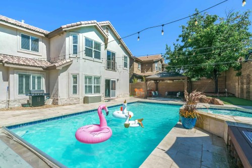 A backyard pool with pink flamingo and white swan floats, surrounded by a house and greenery under a clear blue sky.