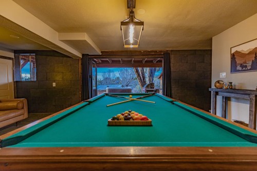 A pool table with balls arranged on it, set in a cozy room with a view of nature through large windows.