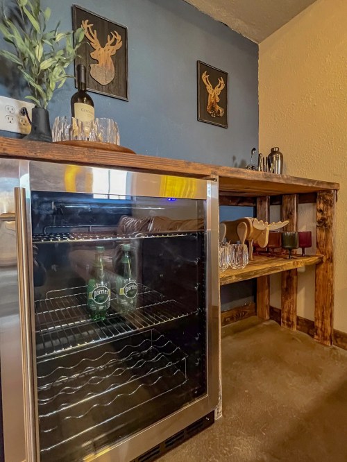 A wooden bar with a wine cooler, glasses, and decorative wall art featuring deer heads.
