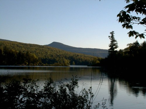 Serene lake surrounded by lush green hills under a clear blue sky, reflecting the landscape in calm waters.