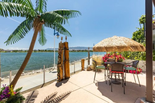 A lakeside patio with a thatched umbrella, chairs, and palm trees, overlooking a serene blue water view.