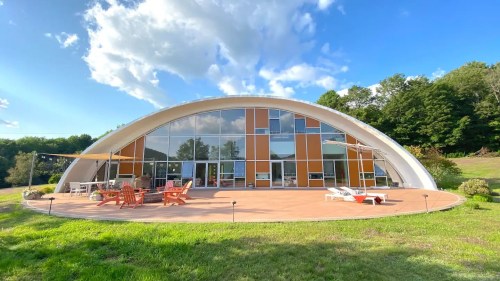 A modern, curved house with large windows, surrounded by green grass and trees under a blue sky with clouds.