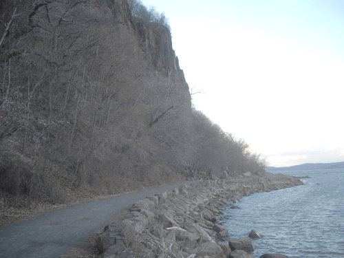 A rocky shoreline with a path alongside a calm body of water, framed by bare trees and a steep cliff in the background.
