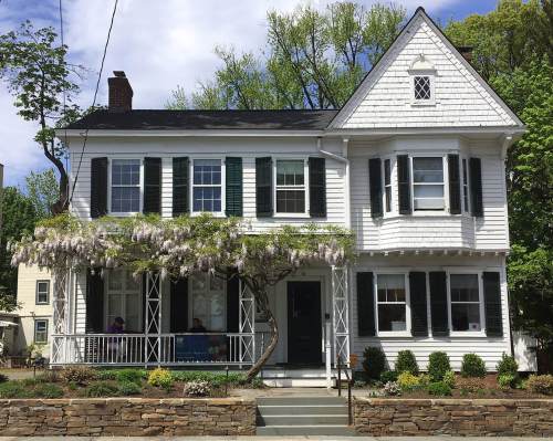 A charming white house with green shutters, featuring a porch and wisteria vines, surrounded by greenery.