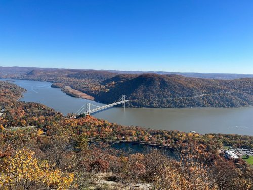A scenic view of a river valley with a bridge, surrounded by autumn foliage and rolling hills under a clear blue sky.