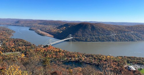 A scenic view of a river valley with a bridge, surrounded by autumn foliage and rolling hills.