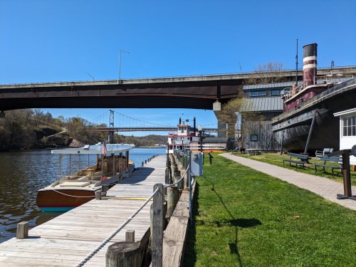 A scenic view of a river with boats docked, a bridge overhead, and green grass along the waterfront.