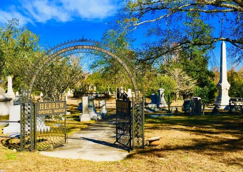 A wrought iron gate labeled "Hebrew Cemetery" opens to a serene graveyard with tombstones under a clear blue sky.