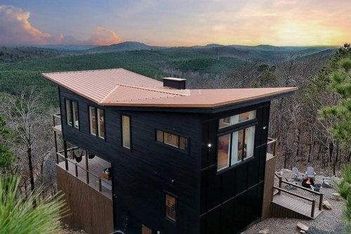 Modern black house with a unique roof design, surrounded by trees and mountains at sunset.